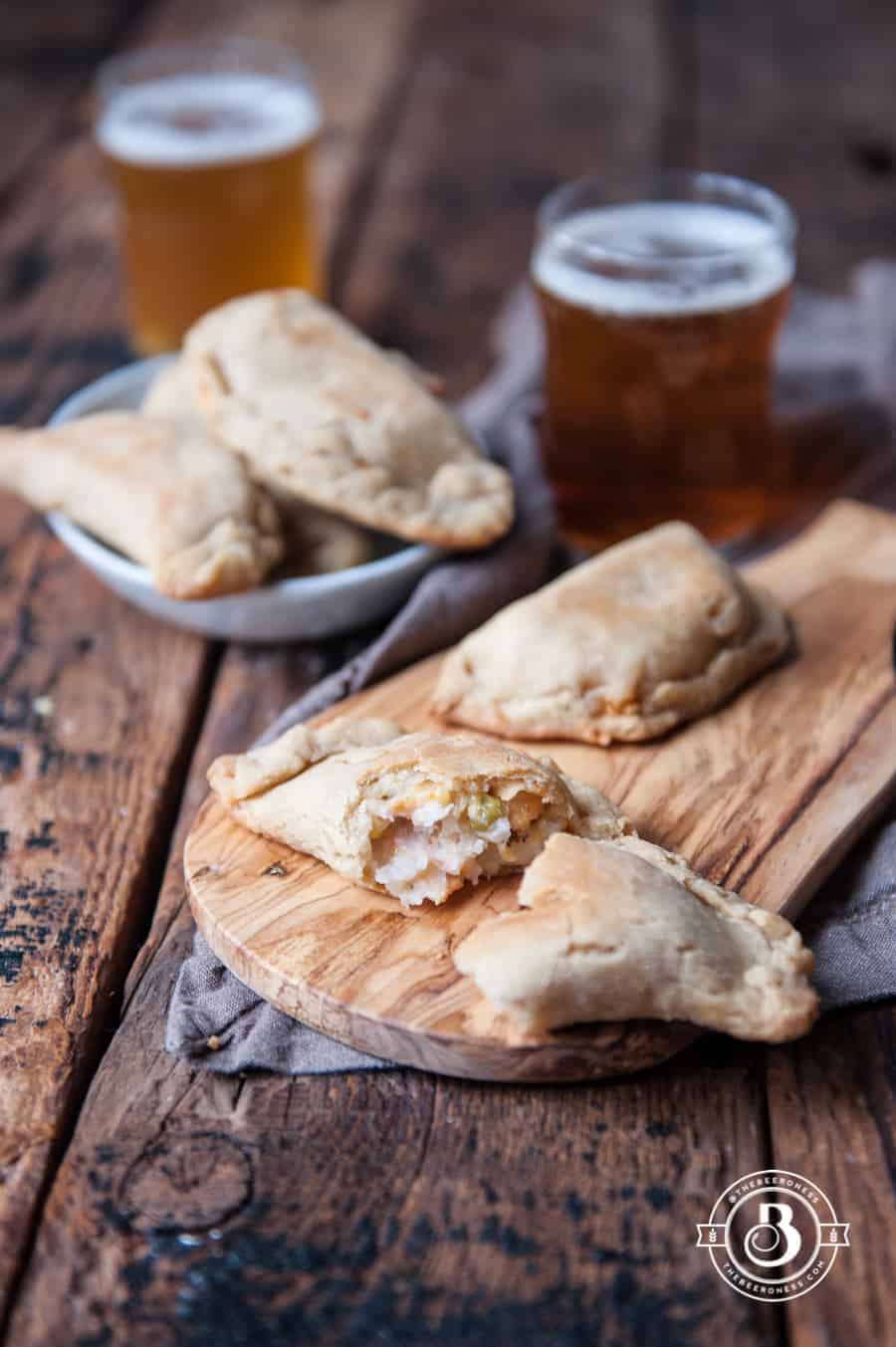 Two turkey empanadas on a wood cutting board by a glass of beer.