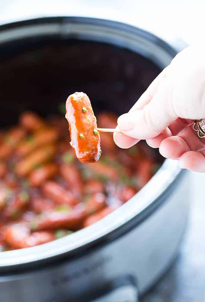 A closeup of a hand holding a little smokie on a toothpick.  A slow cooker is in the background.