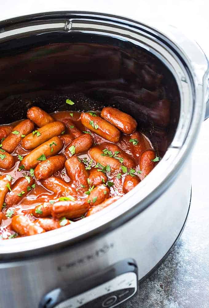 Little smokies in sauce topped with chopped parsley in an oval stainless crockpot.