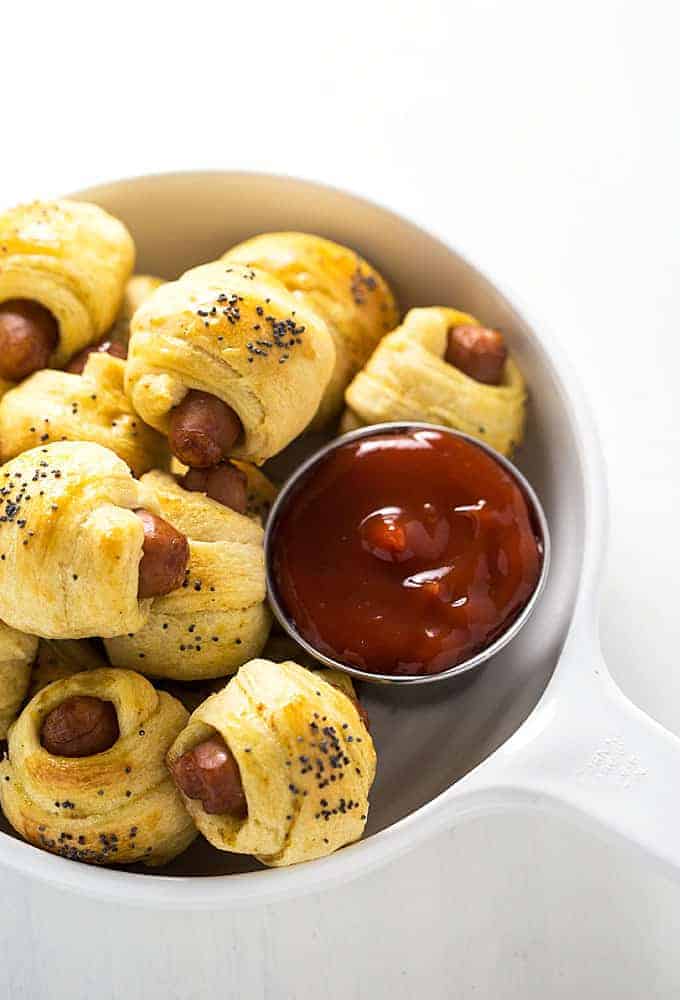 Overhead view of sausages wrapped in crescent rolls with a cup of ketchup in a white bowl.