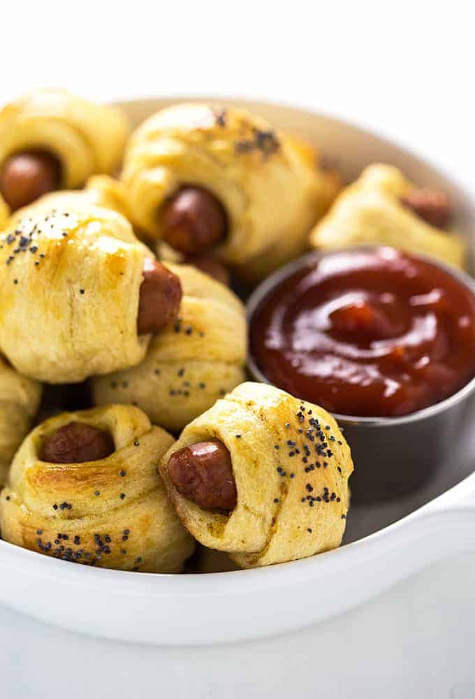 Front closeup view of pigs in a blanket in a white bowl with a small cup of ketchup.