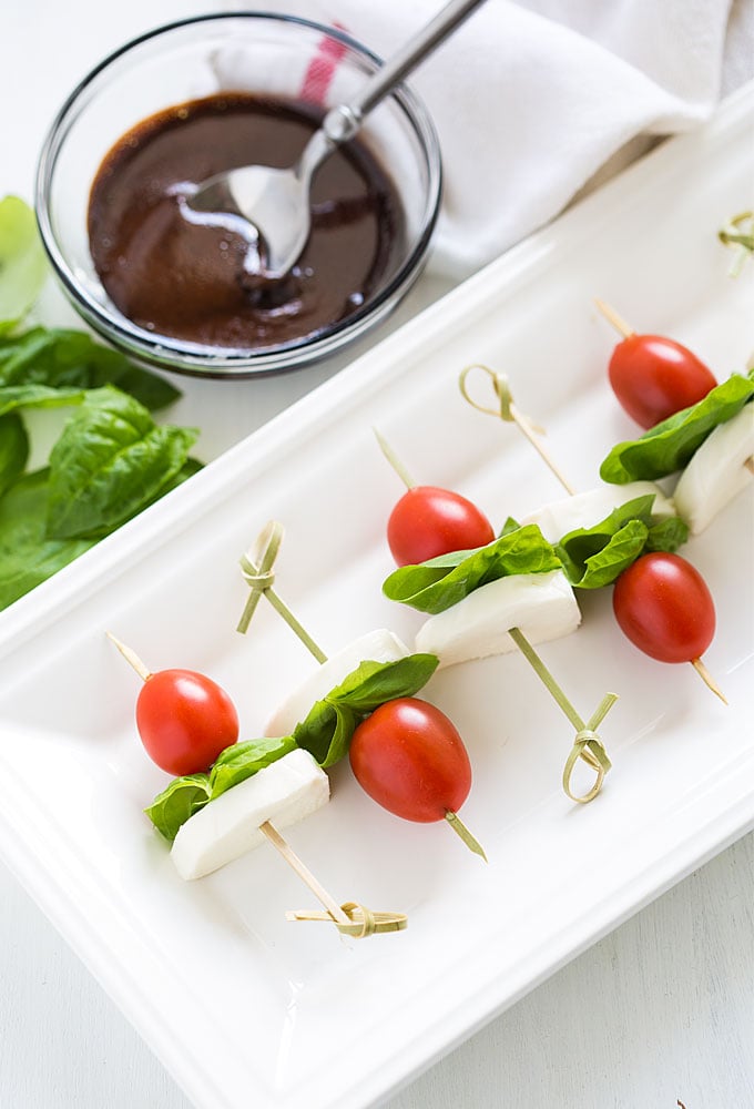 Skewers of tomato, basil and mozzarella on a white serving platter by a bowl of balsamic reduction.