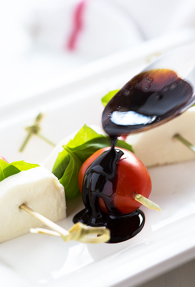 A spoon drizzling a balsamic reduction over a skewer of tomato, basil and mozzarella.