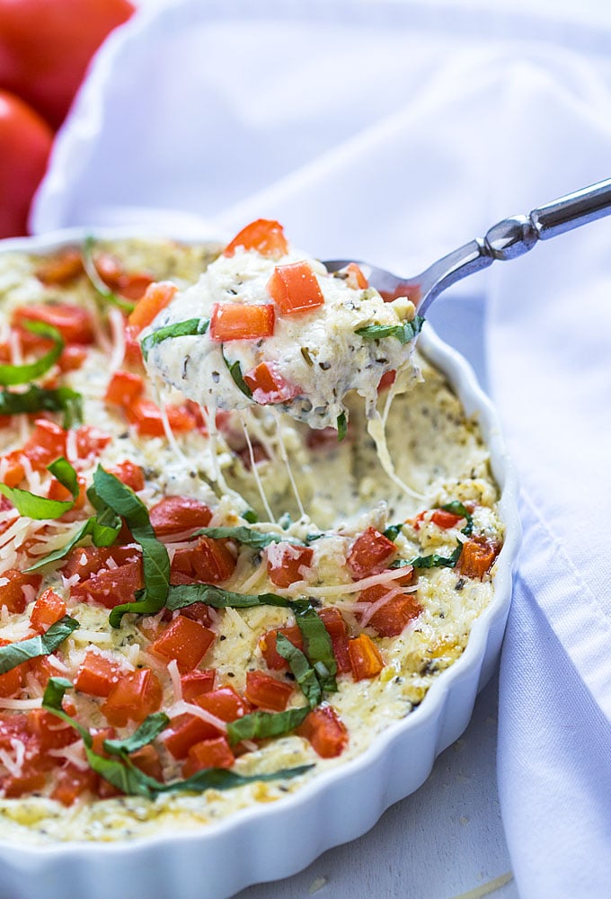 A spoon spooning cheesy caprese dip from a baking dish beside a white napkin.