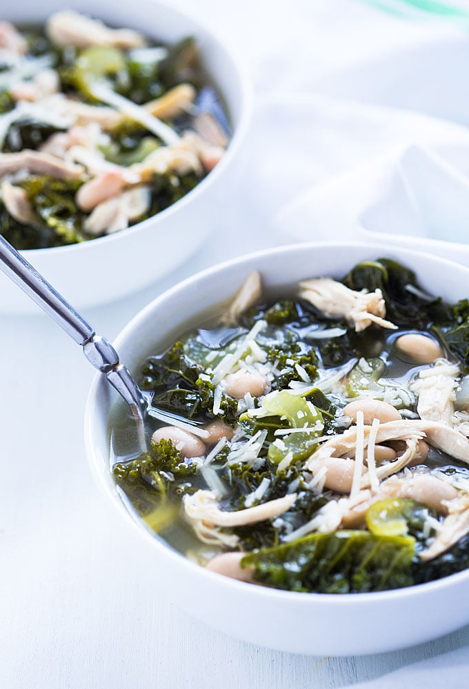 Two white bowls of soup with kale, white beans and chicken topped with grated Parmesan.
