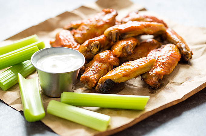 Front view of chicken wings, a cup of ranch dressing and celery sticks on brown paper.