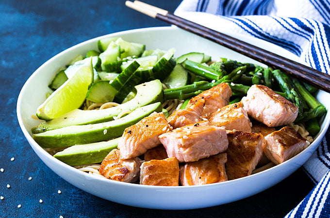 Salmon and noodles with cucumbers, asparagus and avocado in a round white bowl with chopsticks