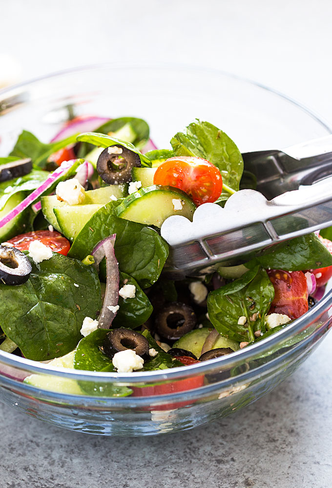 Closeup of salad in a clear glass bowl with a pair of tongs in the bowl.