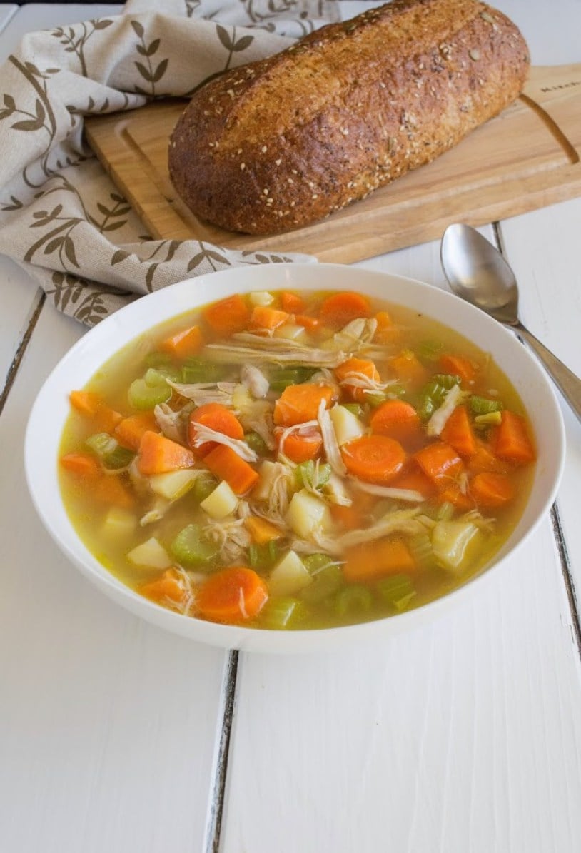 Front view of a white bowl of chicken soup.  A loaf of bread is in the background.