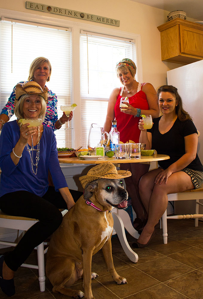 Four women sitting at a table holding margaritas. A boxer dog is sitting on the floor.