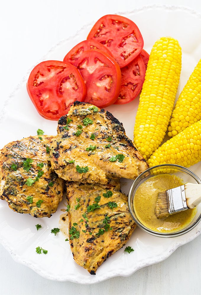 Overhead view of grilled chicken on a platter with a bowl of marinade, corn and tomatoes.