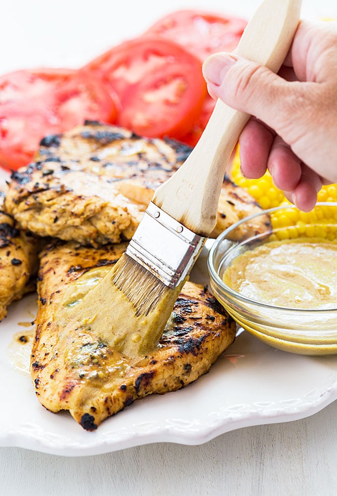 A hand holding a basting brush brushing marinade on a piece of grilled chicken.