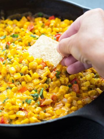 A tortilla chip being dipped into hot corn dip in a cast iron skillet.