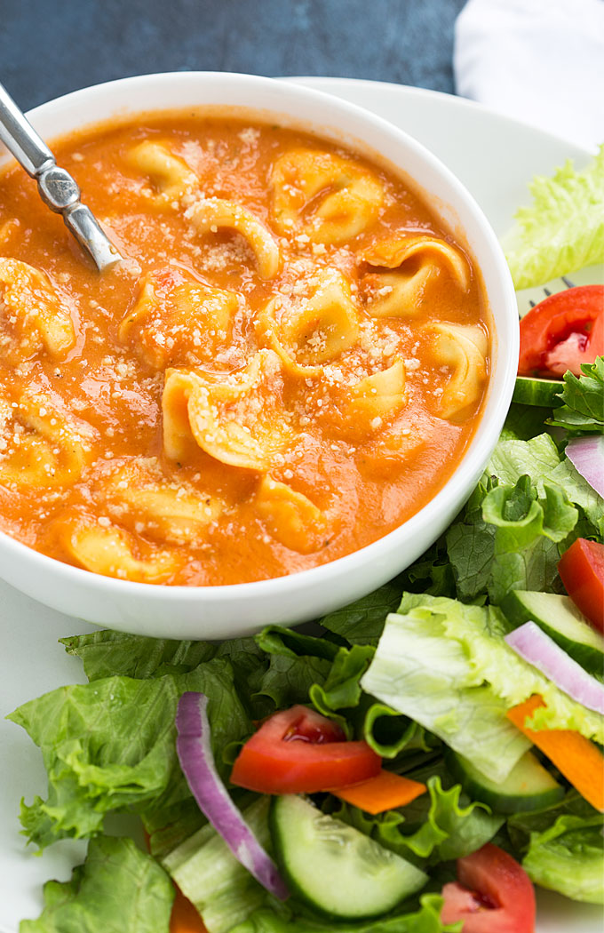 A closeup of a bowl of tomato tortellini soup by a salad.