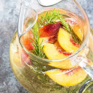 An angled overhead view of white sangria with peaches and rosemary in a pitcher.