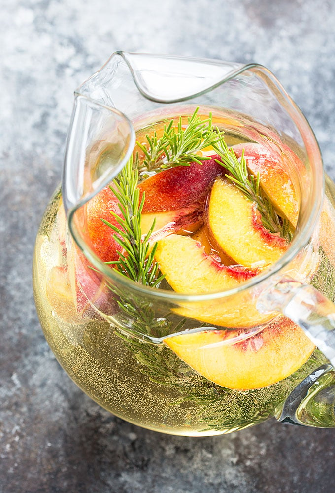Overhead view of sangria with white wine, fresh peaches and rosemary in a glass pitcher.