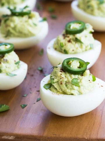A closeup of guacamole deviled eggs topped with sliced serrano peppers on a wood cutting board.
