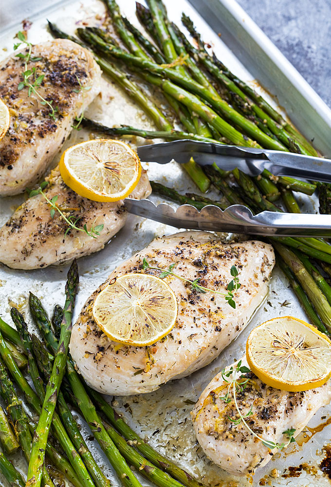 Baked chicken with lemon and asparagus on a baking sheet with a pair of tongs.