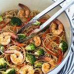 Shrimp lo mein with broccoli in a white dutch oven with a pair of tongs.