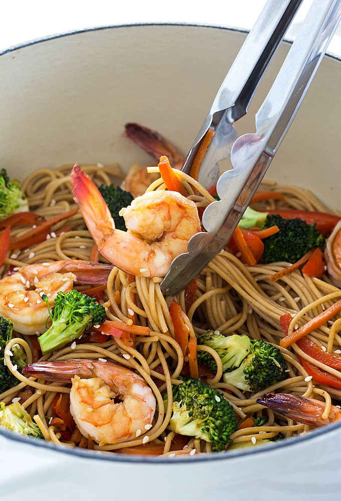 A closeup of a pair of tongs removing lo mein from a white dutch oven.