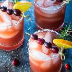 Three margaritas garnished with cranberries, orange and rosemary on a dark surface.