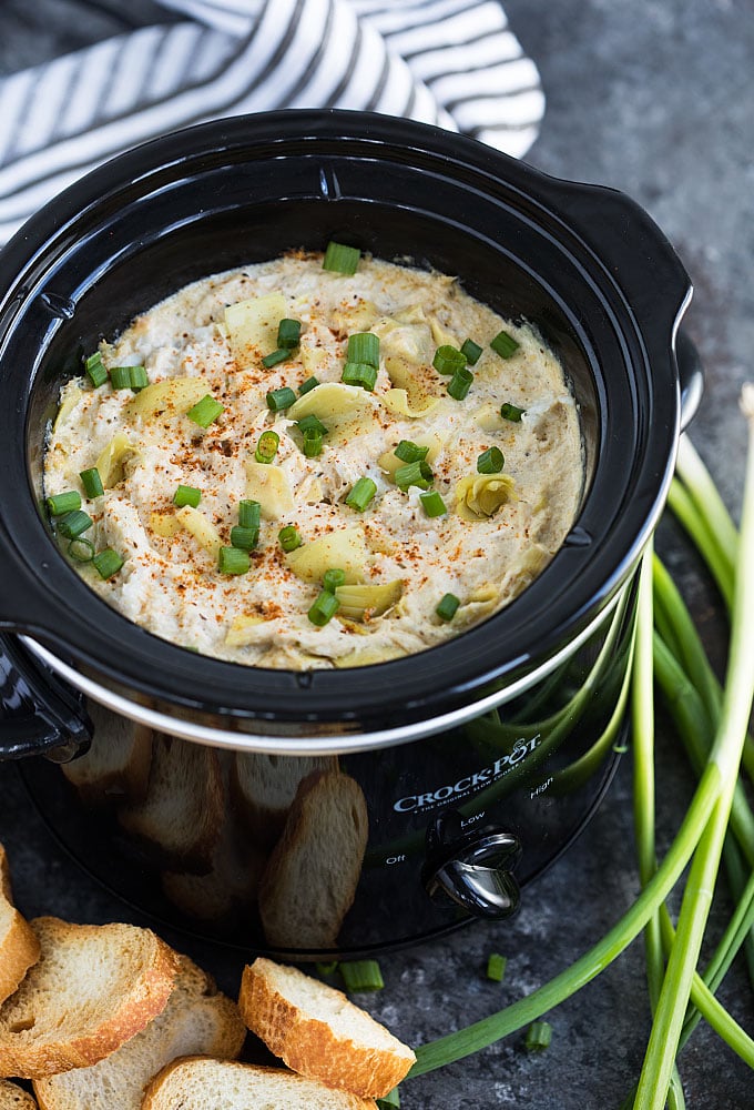 Crab and artichoke dip with green onions in a round black slow cooker.