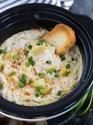A piece of French bread dipped into artichoke crab dip in a slow cooker.