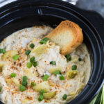 Crab and artichoke dip in a round black crock pot with overlay text.