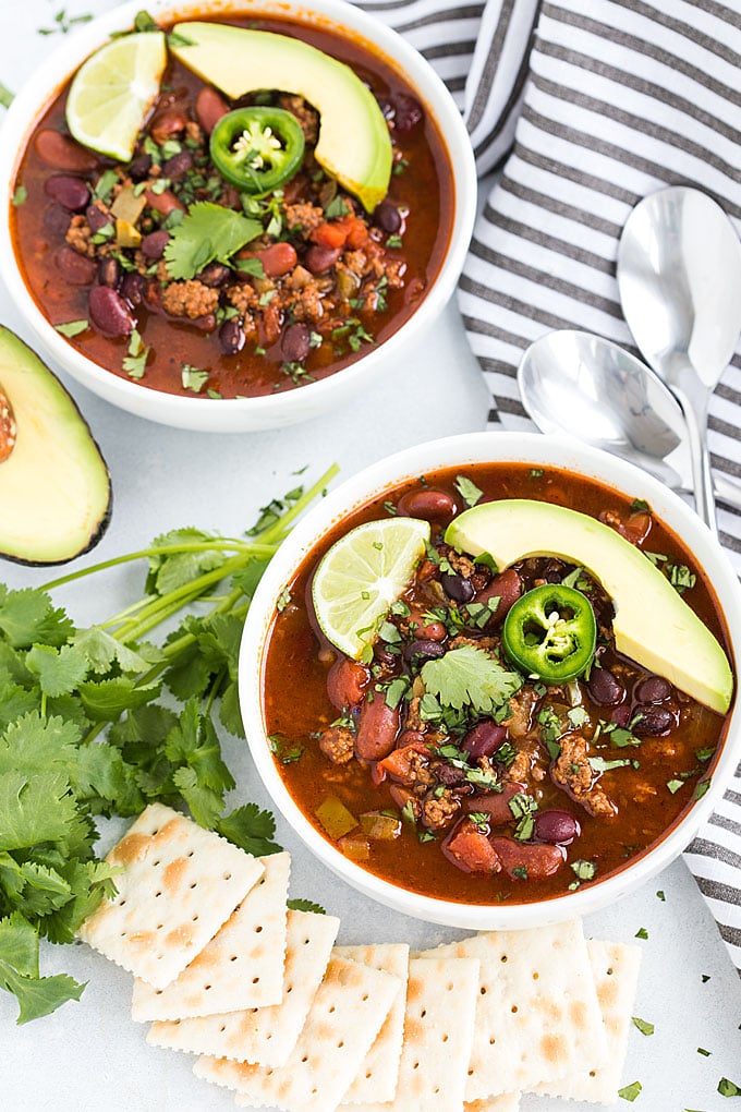 Overhead view of two white bowls of chili topped with avocado, jalapeno, cilantro and lime.