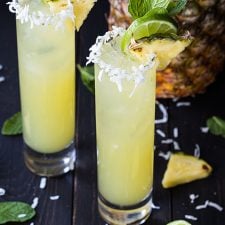 Two mojitos garnished with pineapple, lime and mint in glasses rimmed with flaked coconut.