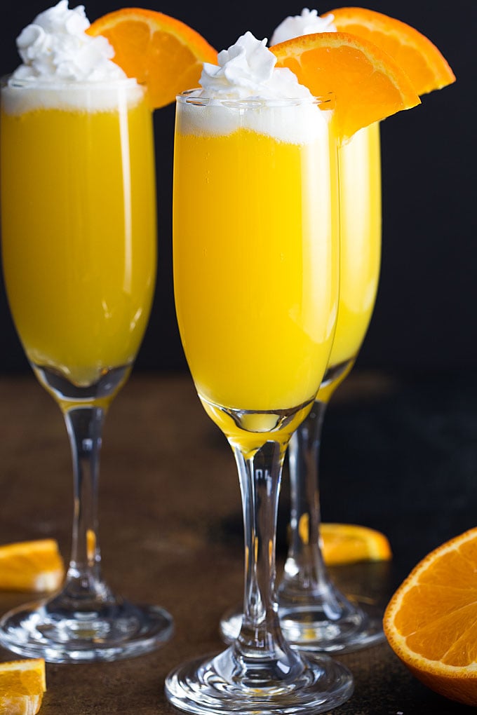 Front view of three mimosas with whipped topping and an orange slice on a dark surface.