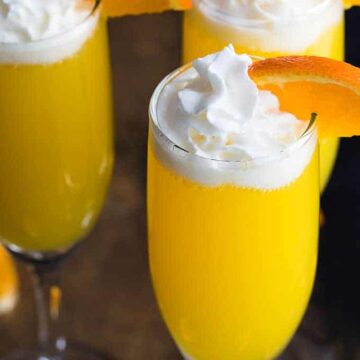 Three creamsicle mimosas with whipped topping and an orange slice in champagne flutes.