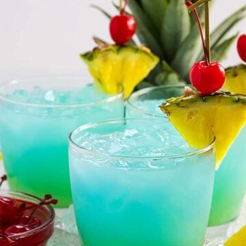 Front view of three blue and green cocktails garnished with pineapple and cherries.
