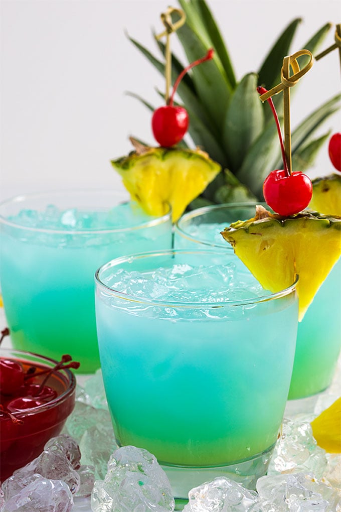 Front view of three blue and green cocktails garnished with pineapple and cherries.