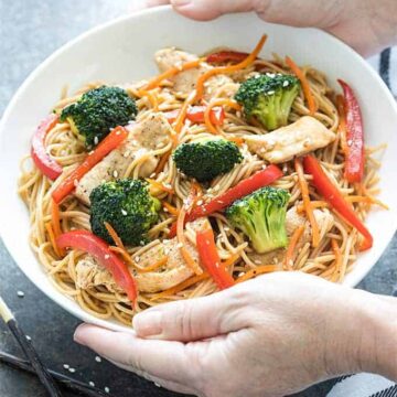 Two hands holding a white bowl of chicken and broccoli lo mein over a dark surface.