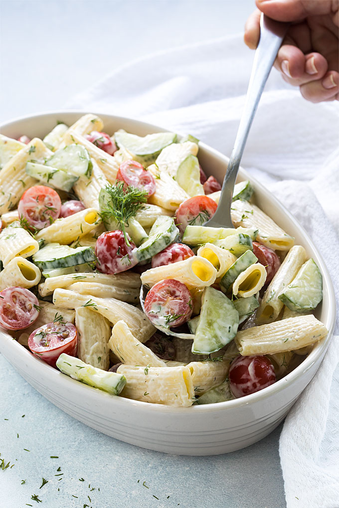 A spoon spooning a serving of creamy cucumber and tomato pasta salad from an oval dish.