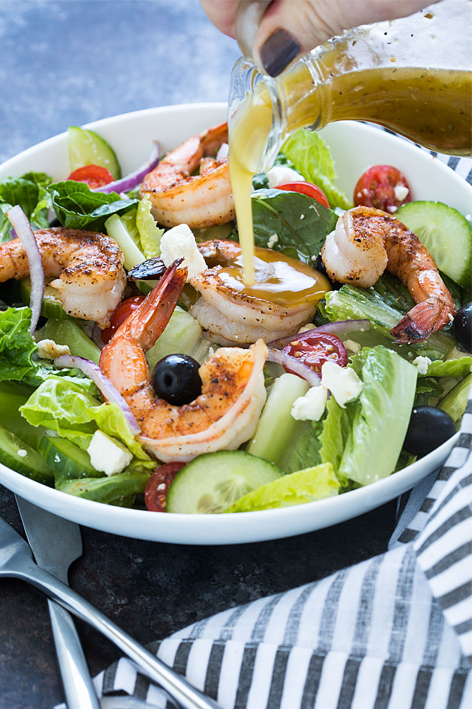 Dressing being poured over a Greek salad with blackened shrimp in a white bowl.