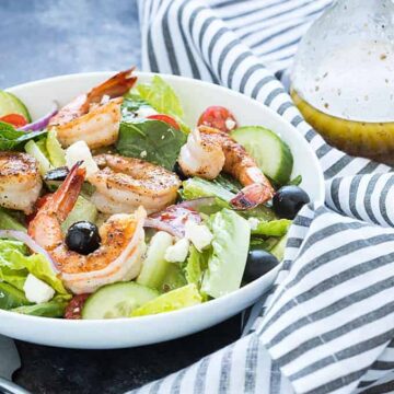 A white bowl of salad with shrimp beside a bottle of Greek dressing and a napkin.