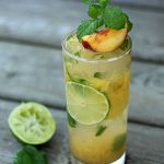 A mojito in a glass garnished with a peach slice and mint on a wood surface.