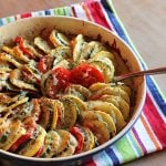 Baked thinly sliced squash, tomatoes, potatoes topped with cheese in a baking dish with a spoon.
