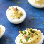 Deviled eggs with bacon and topped with chopped parsley on a blue surface.
