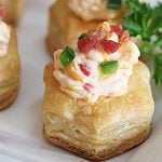 A closeup of a puff pastry cup filled with pimento cheese and sprinkled with bacon pieces.