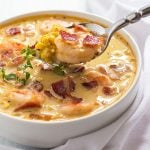 A spoon removing soup from a white bowl of bacon, shrimp and corn chowder.