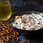 A gray bowl of dip by pretzels and a glass of beer.