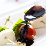 A spoon drizzling balsamic reduction on a tomato on a skewer with mozzarella and basil.