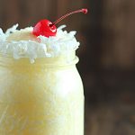 An orange frozen cocktail with a cherry in a jar rimmed with shredded coconut.