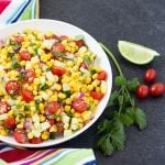A salad with corn, tomatoes and cucumbers in a white bowl by a striped towel.