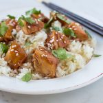 Bite size chicken pieces with orange sauce over rice on a white plate with chopsticks.