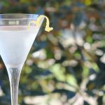 Front view of a martini garnished with lemon peel.  A bush is in the background.