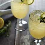 Two iced lemonade cocktails garnished with lemon and fresh thyme on a wood surface.